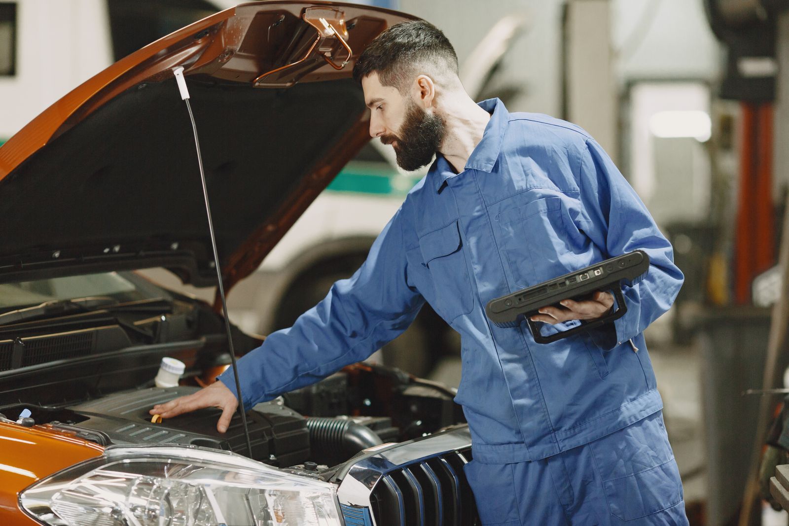 Mechanic in blue coveralls standing beside an open car bonnet, holding a handheld diagnostic tablet, looking into the engine bay