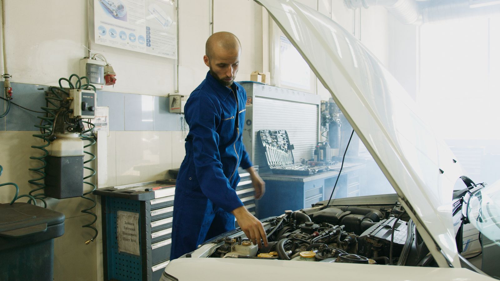 Mechanic in blue coveralls working on an engine bay of a white vehicle in a workshop bay with tool cabinet and pegboard visible behind
