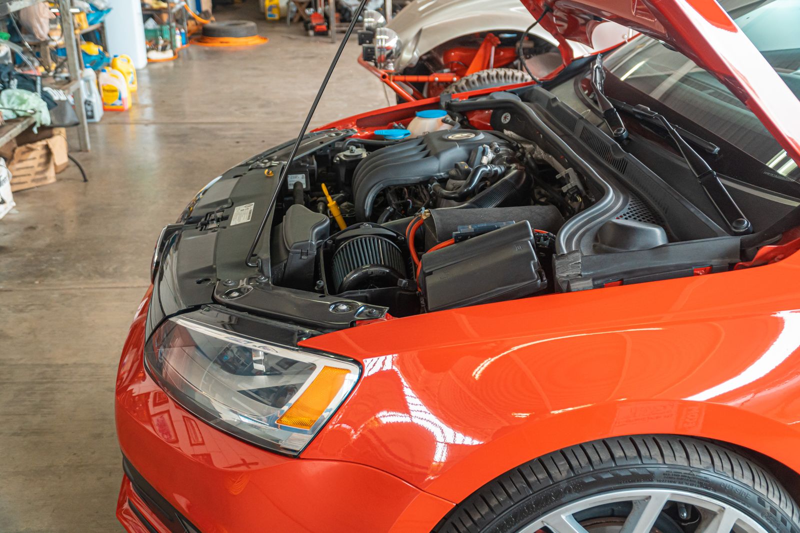 Close-up of an open engine bay on a red hatchback with aftermarket intake visible inside a workshop