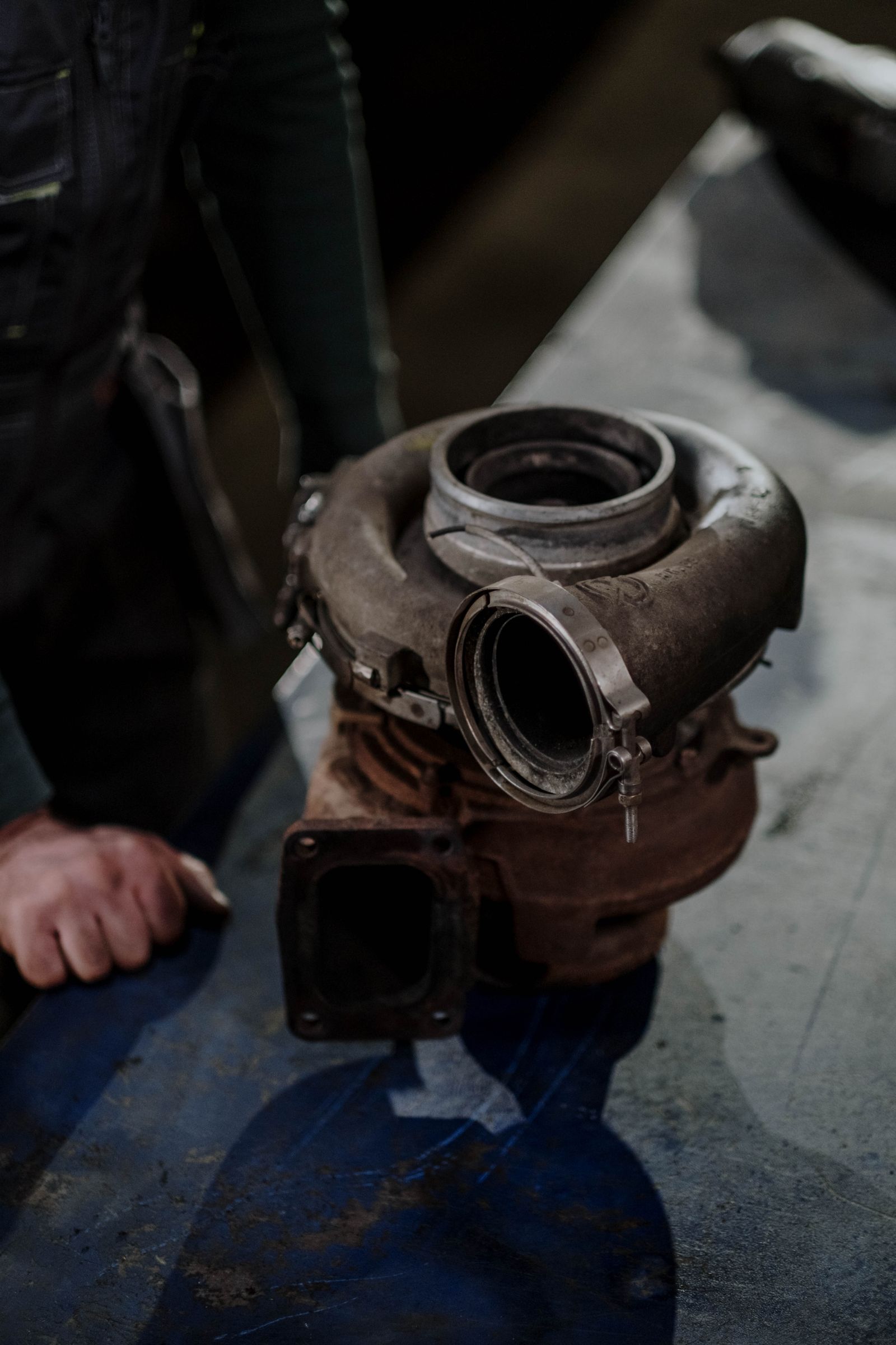 Close-up of a removed turbocharger unit sitting on a workshop bench with a mechanic's hand visible in the background