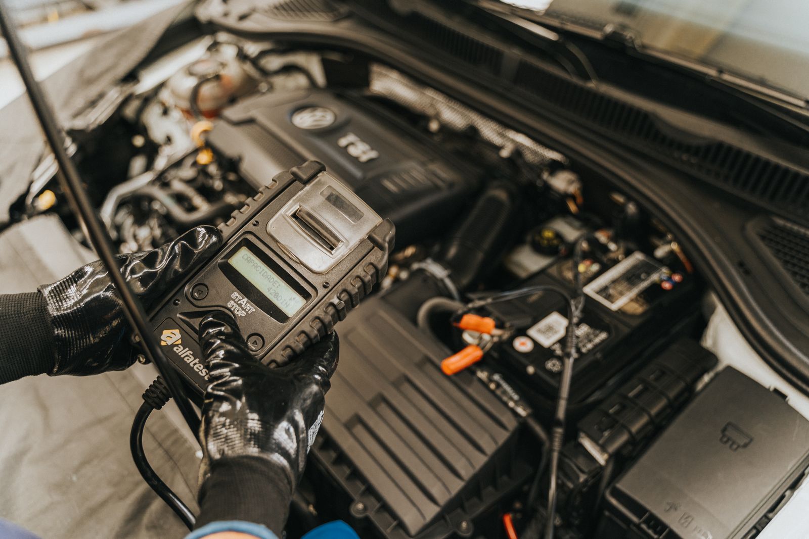 Gloved hands holding a handheld diagnostic scan tool up to a Volkswagen engine bay with VW roundel visible on the engine cover